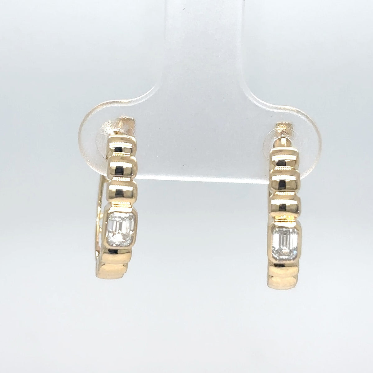 A pair of 14K yellow gold hoop earrings with emerald-cut diamonds, displayed on a clear stand against a light gray background.
