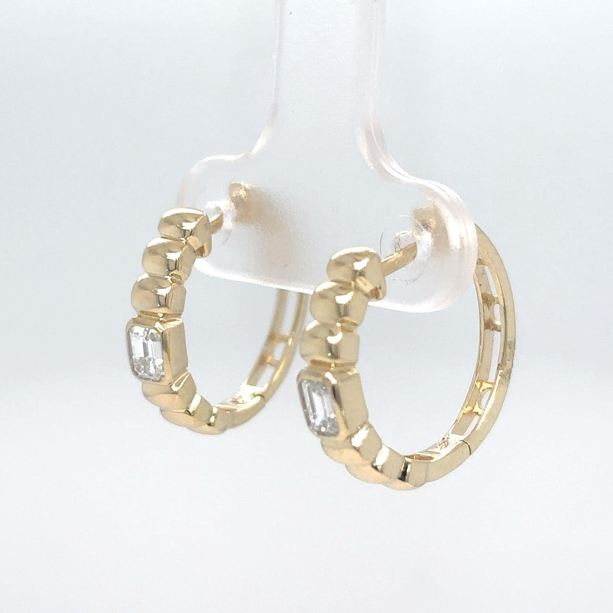 A pair of 14K yellow gold hoop earrings with textured beaded edges and emerald-cut diamonds, displayed on a clear hanger against a white background.