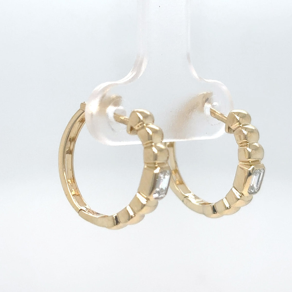 A pair of 14K yellow gold beaded hoop earrings, each set with an emerald-cut diamond, displayed on a clear stand against a white background.