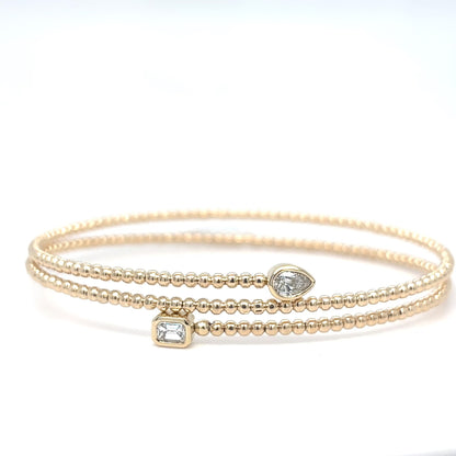 A delicate 14K yellow gold beaded flex bangle bracelet with a bezel-set emerald-cut diamond and a pear-shaped diamond accent, displayed on a white background.