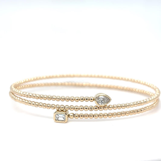 A delicate 14K yellow gold beaded flex bangle bracelet with a bezel-set emerald-cut diamond and a pear-shaped diamond accent, displayed on a white background.