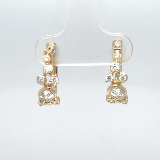 A pair of 14K yellow gold huggie hoop earrings set with emerald-cut and round brilliant diamonds, shown on a clear display stand against a white background.