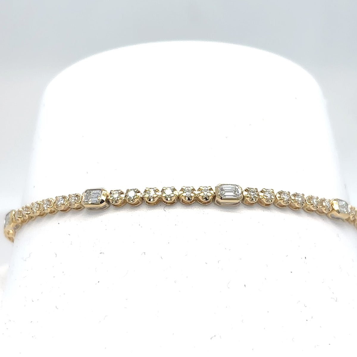 An 18K yellow gold tennis bracelet set with emerald-cut and round brilliant diamonds, displayed on a white cylindrical stand against a light gray background.