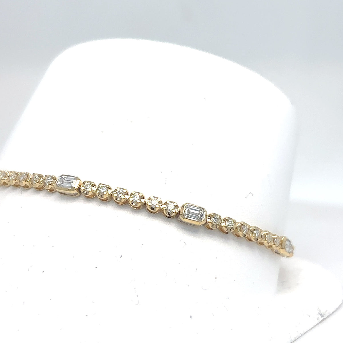 An 18K yellow gold tennis bracelet with emerald-cut and round brilliant diamonds, displayed on a white jewelry bust.