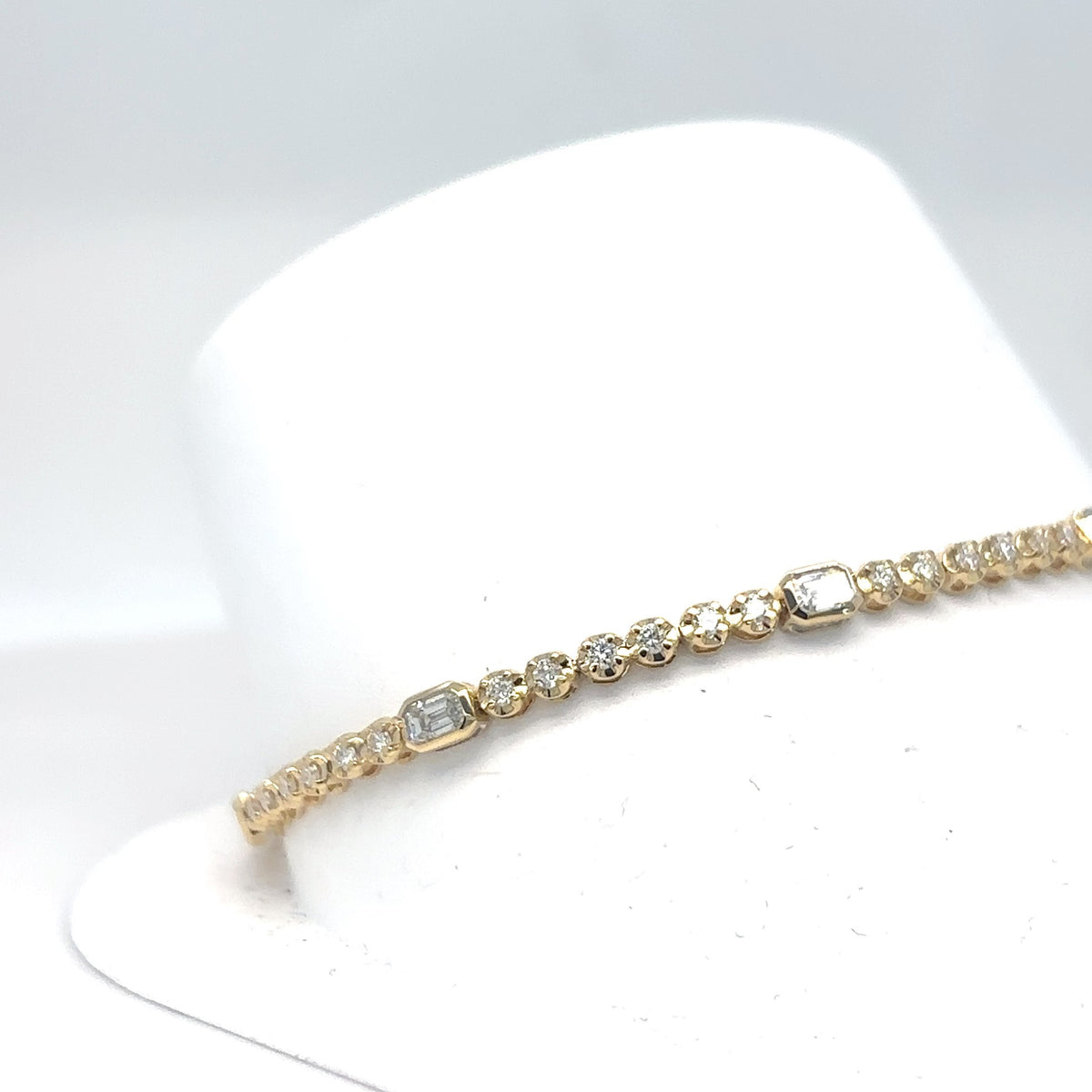 An 18K yellow gold tennis bracelet with alternating emerald-cut and round brilliant diamonds, displayed on a white jewelry stand against a light gray background.
